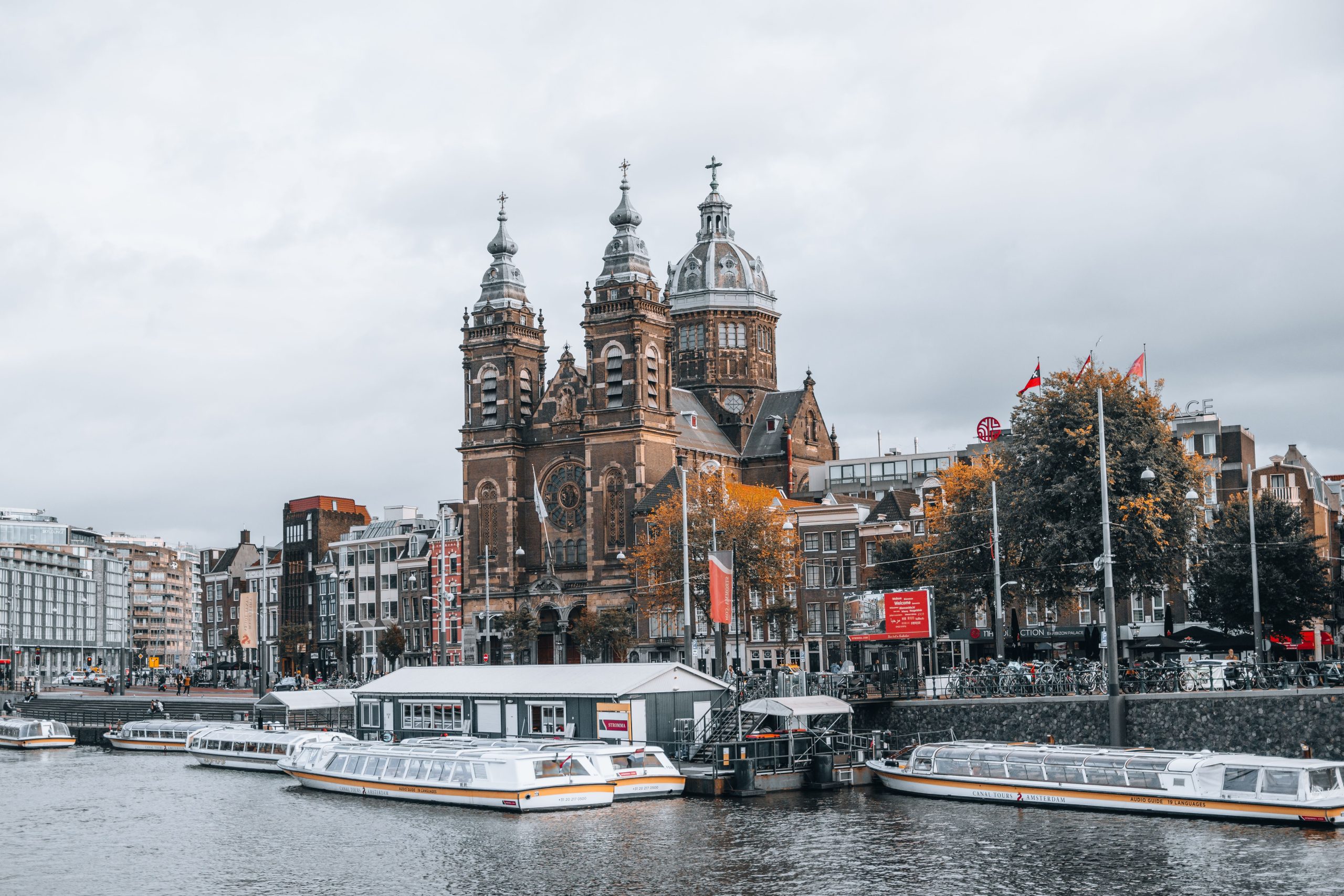 Historic church with towers by waterfront, boats docked in foreground, cloudy sky.